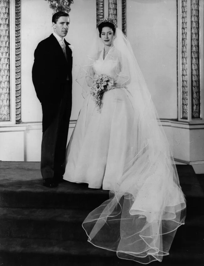 Elegant couple on wedding day, bride in a long gown and veil with bouquet; groom in a suit, standing on carpeted stairs.