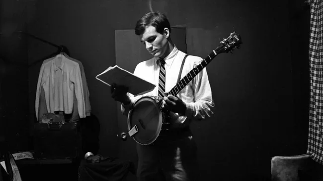 Black and white photo of a man in a tie holding a banjo, reading sheet music in a dimly lit room.