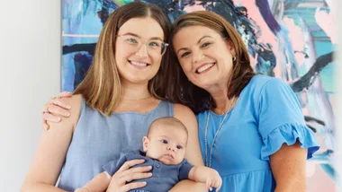 Two women smile, standing closely, with one holding a baby. A colorful abstract painting is in the background.