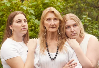 Three women standing close together, looking serious, outdoors with greenery in the background.