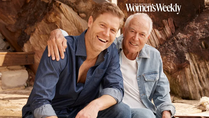 Two men smiling, sitting close together outdoors, wearing casual denim shirts. One has his arm around the other.