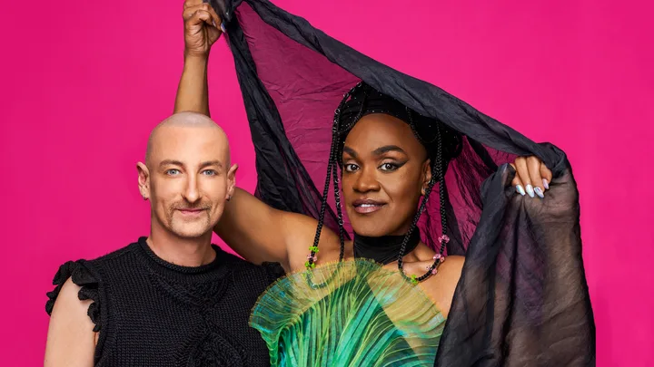 Two individuals pose against a pink background, one in a black sleeveless top, the other in a vibrant green outfit holding fabric.