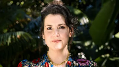 A woman with dark hair in a colorful blouse stands outdoors against a blurred green foliage background.