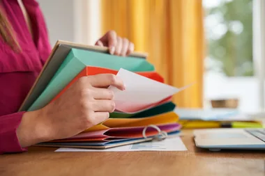 Person sorting papers in a colorful expandable file folder on a wooden desk beside a laptop.