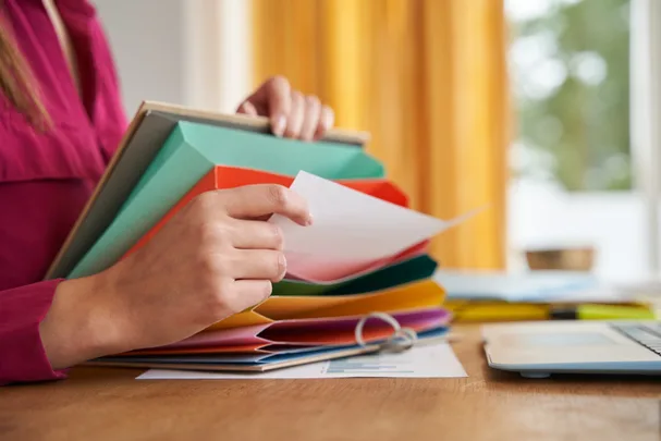 Person sorting papers in a colorful expandable file folder on a wooden desk beside a laptop.