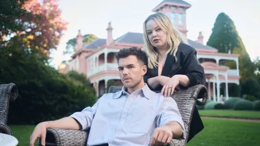 A man seated and a woman standing behind him in front of a Victorian-style house.