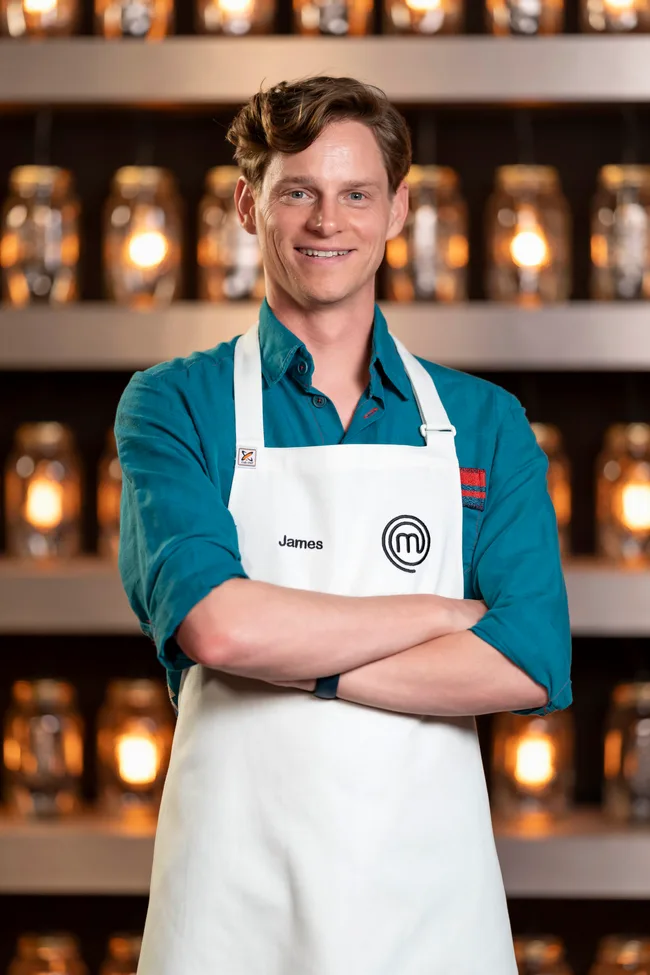 Person named James in a white cooking apron, smiling with crossed arms in front of blurred shelves with lit jars.