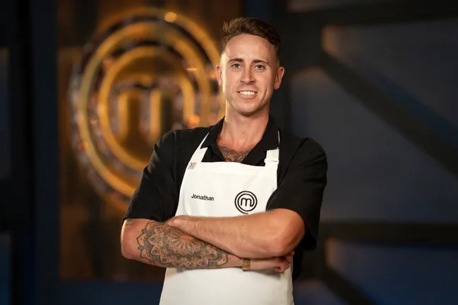 A man named Jonathan in a MasterChef apron stands confidently with crossed arms in front of a MasterChef logo.