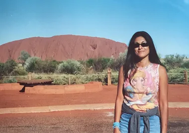Woman in sunglasses stands in front of Uluru with a clear blue sky.