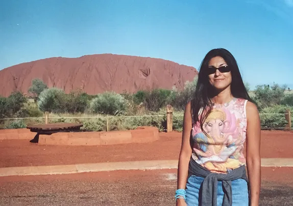 Woman in sunglasses stands in front of Uluru with a clear blue sky.