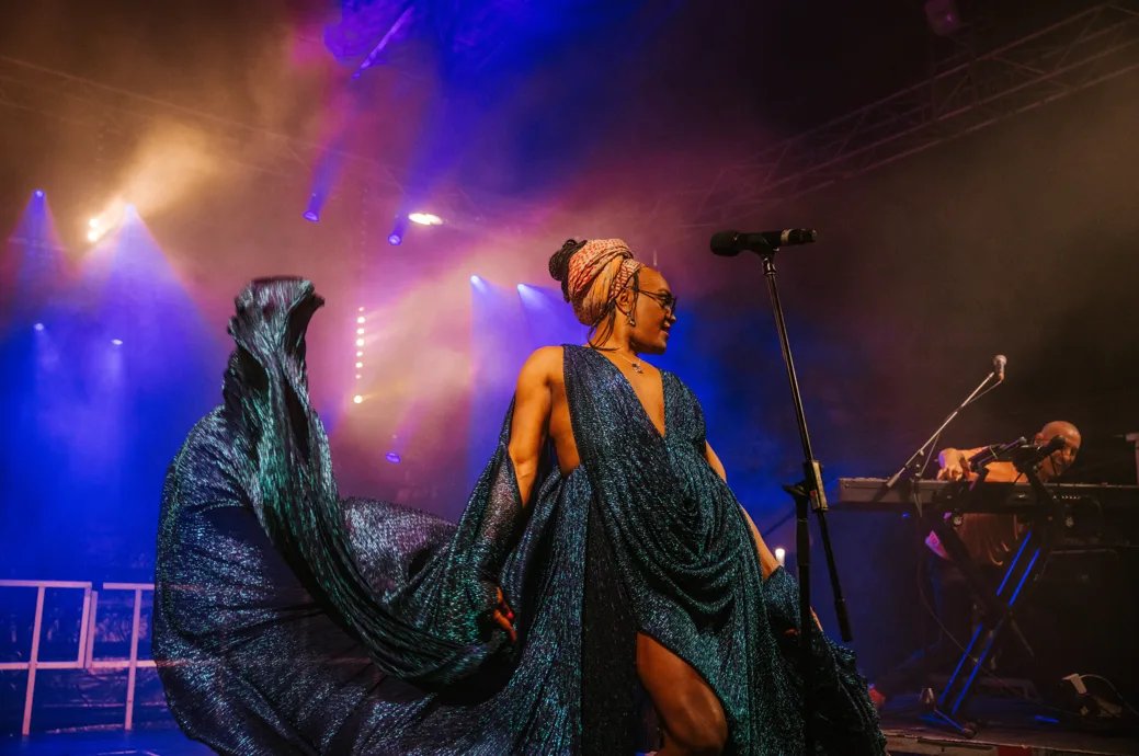 Zaachariaha performs live on stage wearing a sparkly blue dress and shawl and an earth-toned turban.