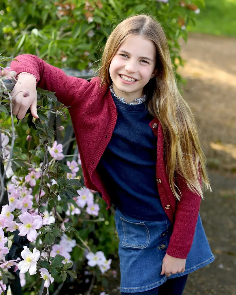 Girl with long hair in red cardigan and denim skirt smiles, leaning on a garden fence with pink flowers.