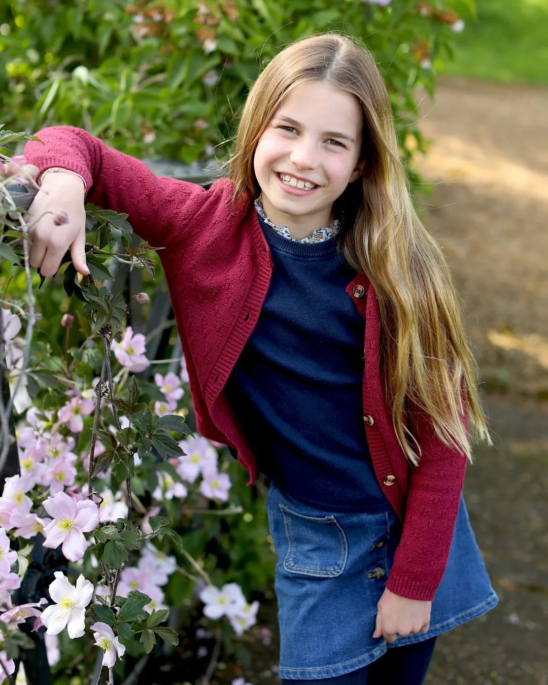 Girl with long hair in red cardigan and denim skirt smiles, leaning on a garden fence with pink flowers.