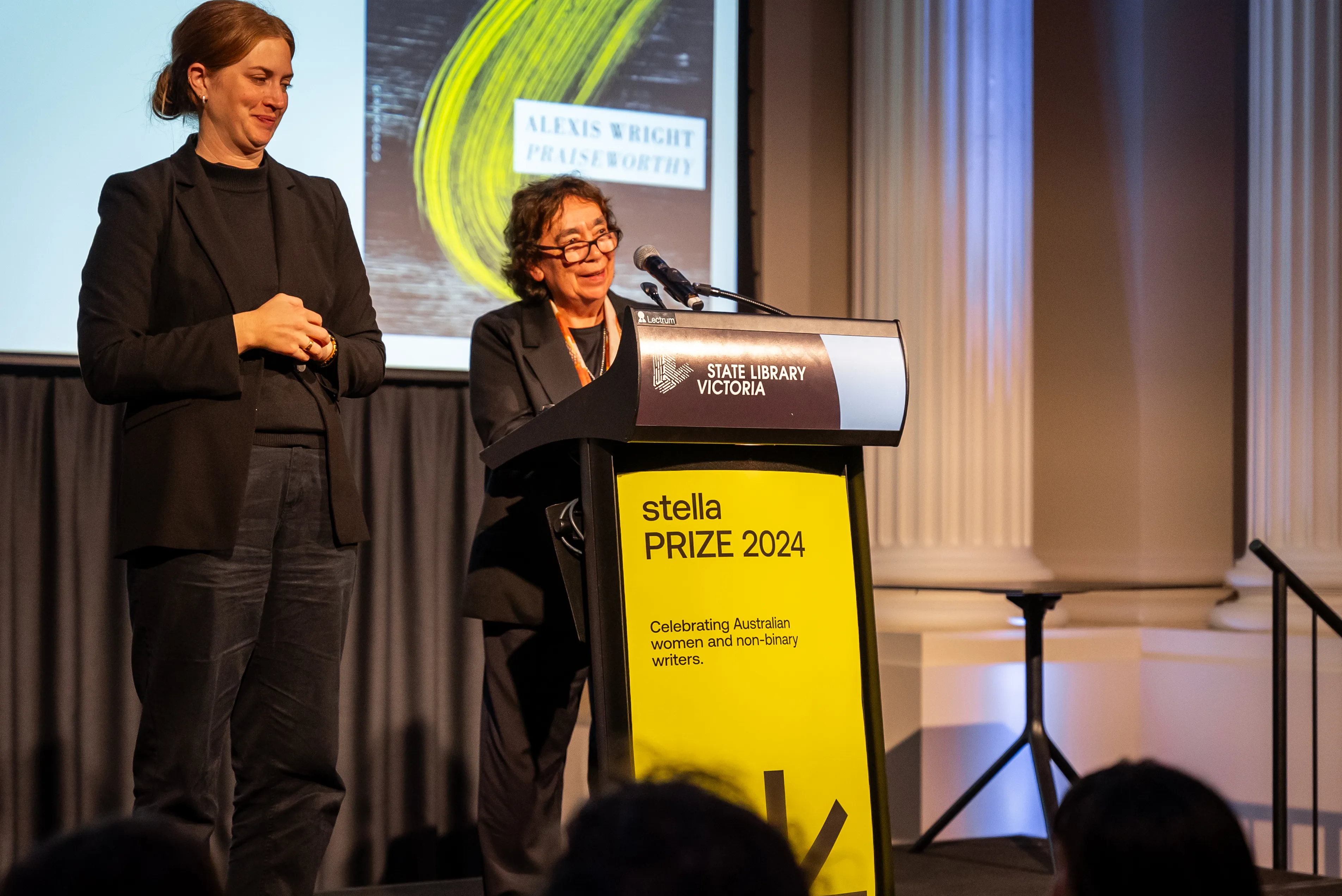 Alexis Wright stands on the stage smiling as she makes her acceptance speech. The AUSLAN interpreter stands chuckling at her side. Both women wear black suits.