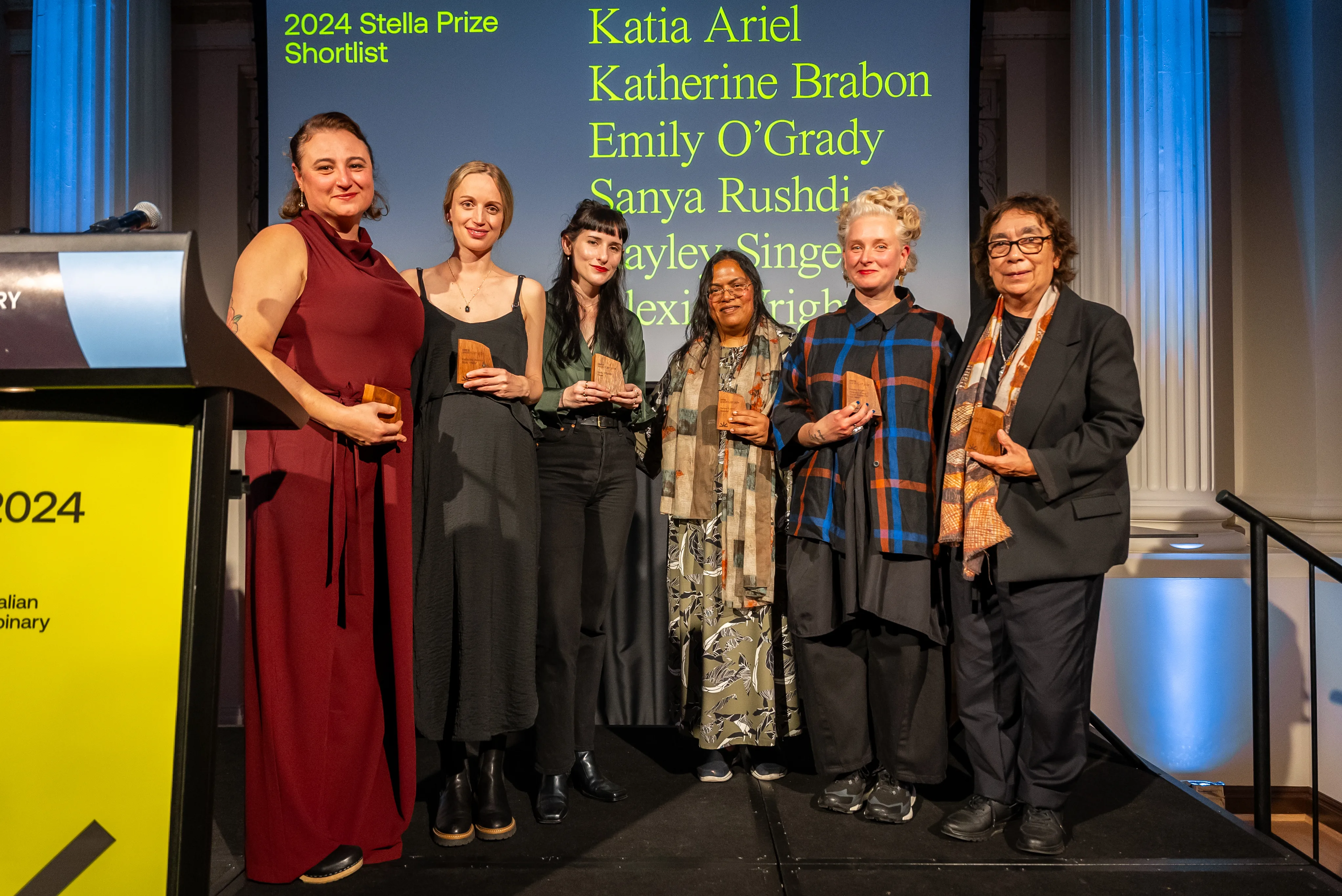 Alexis Wright, wearing a black suit and an earth-toned scarf, stands on the stage with other Stella Prize nominees.