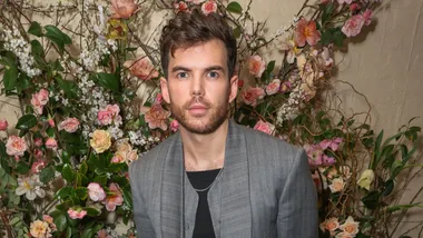 A person in a gray jacket stands in front of a floral background with pink and white blossoms.