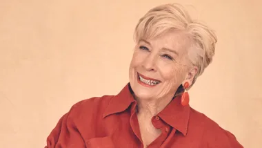 Elderly woman with short blonde hair in red shirt and earrings, smiling against a beige background.