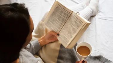 Person reading a book and holding a cup of coffee in bed, wearing cozy attire.