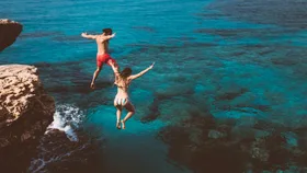 Two people in swimwear jumping off a rocky cliff into the clear blue sea.