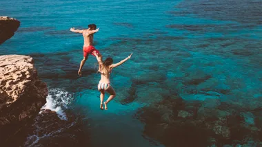 Two people in swimwear jumping off a rocky cliff into the clear blue sea.