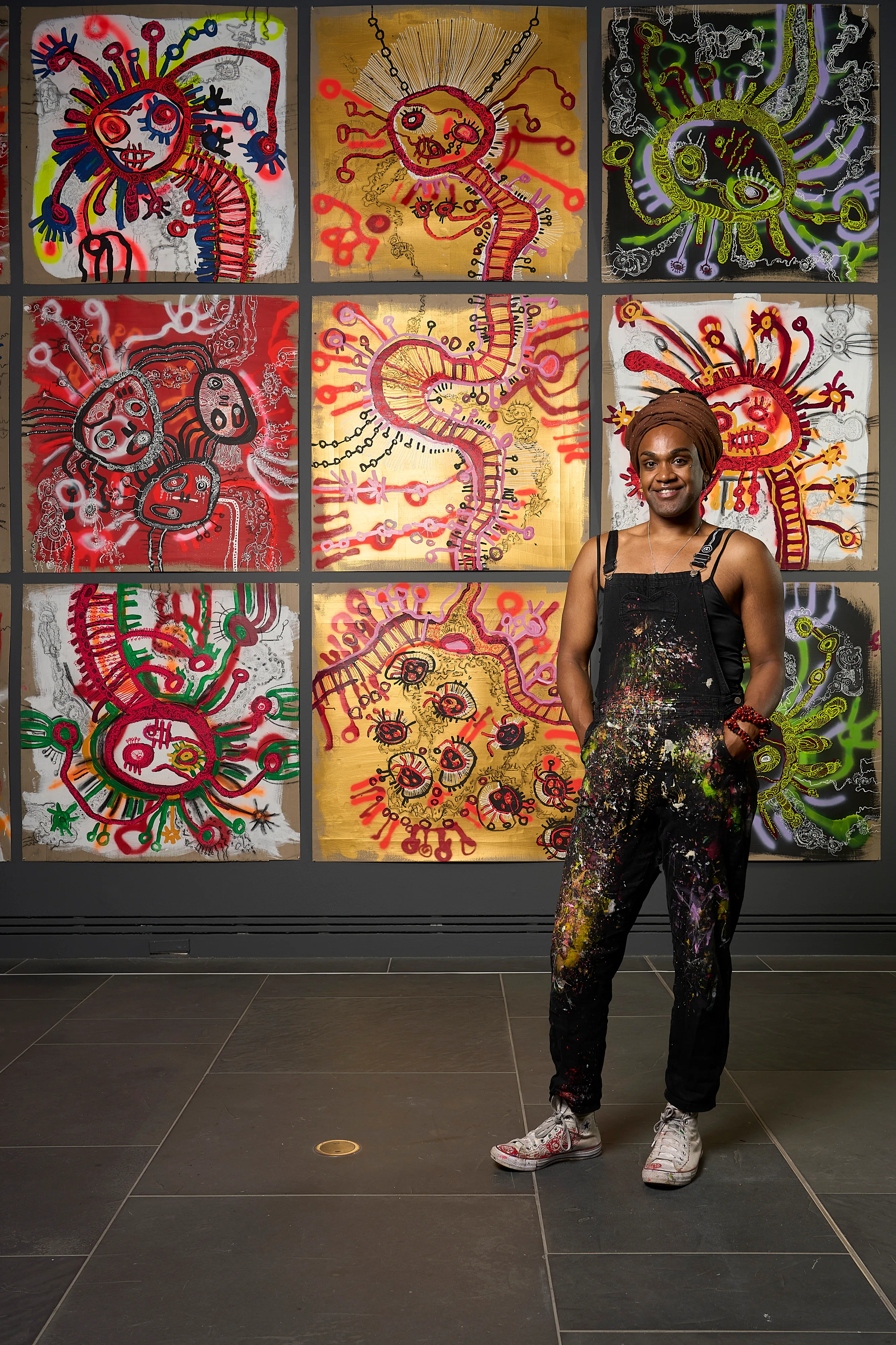 Zaachariaha Fielding wears paint-splattered black overalls and stands in front of a brightly coloured painting.