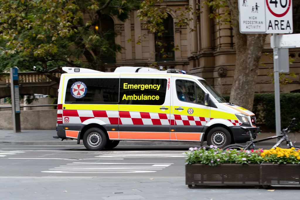 Ambulance parked on a city street, with New South Wales Ambulance logo, next to road signs and trees.