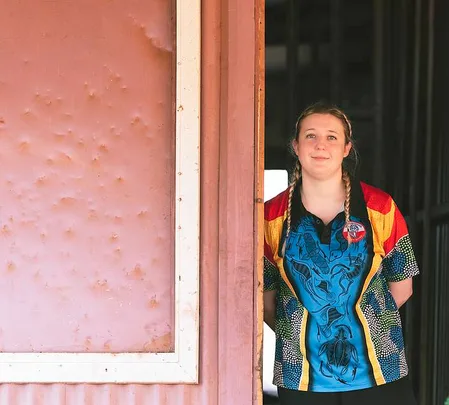 Young woman with braids and colorful shirt standing in a doorway, with a textured wall on the left.