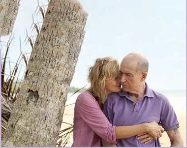 A couple embraces affectionately on a sandy beach, with palm trees and the ocean in the background.