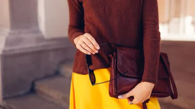 A close-up lifestyle shot of a woman wearing a chocolate brown fine-knit sweater, styled with a vibrant yellow skirt while holding a dark brown leather satchel and black sunglasses.