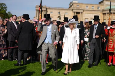 A formal event at Buckingham Palace garden with guests in suits and hats; a man and woman lead the group.