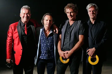 Four men standing together, one in a red jacket, holding awards, smiling at the camera against a dark background.