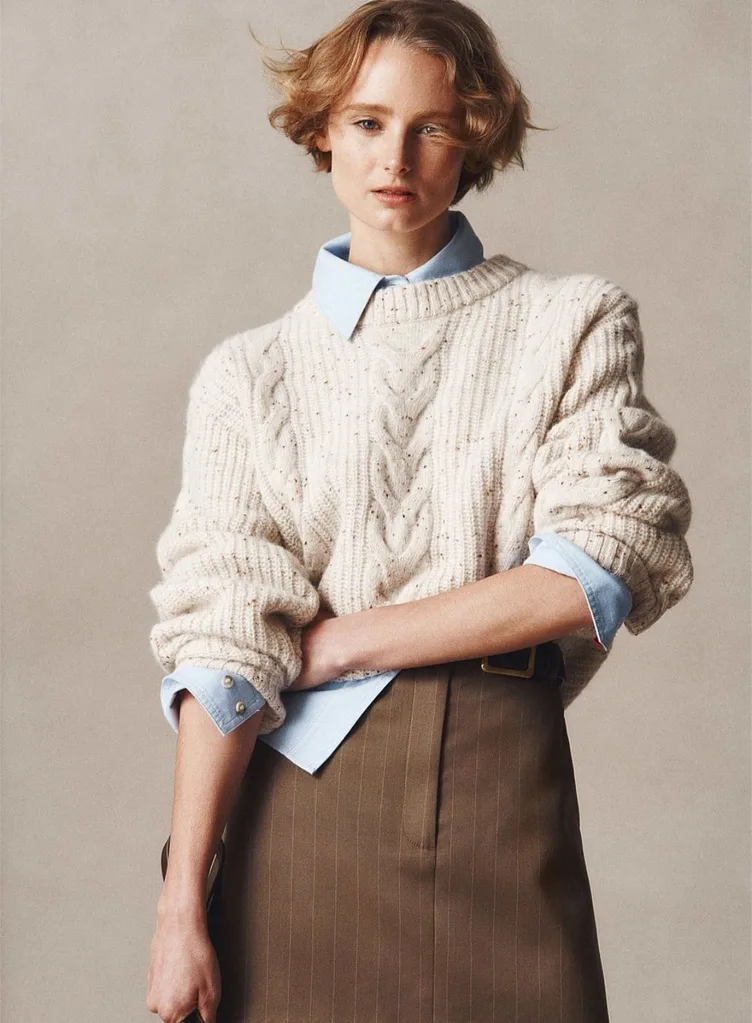 A model with short, wavy auburn hair wearing the Country Road longline cable knit jumper in snow marle. The cream-coloured, chunky knit sweater features a classic cable pattern and is layered over a light blue collared shirt and brown pinstripe trousers.