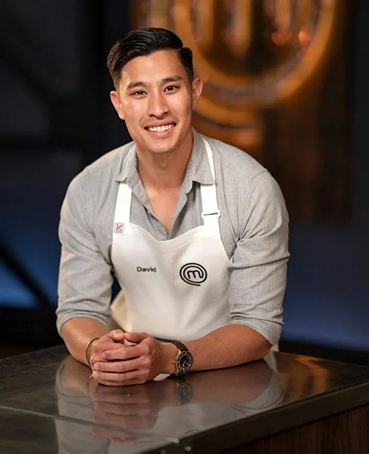 "Smiling chef David wearing a MasterChef apron, leaning on a kitchen counter."