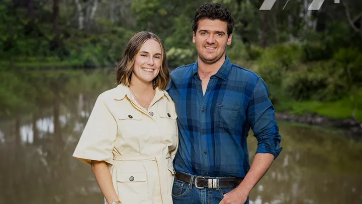 Couple smiling outdoors with trees and a pond in the background.