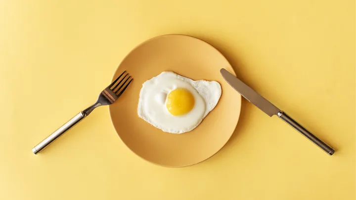 Fried egg on a yellow plate with silver fork and knife on a yellow background.
