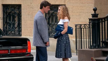 A woman in a white top talks to a man beside a parked car on a city street.