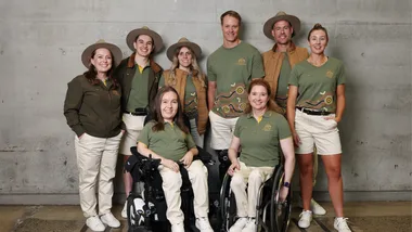 Australian Paralympic team members in uniform, two in wheelchairs, smiling indoors, concrete wall background.