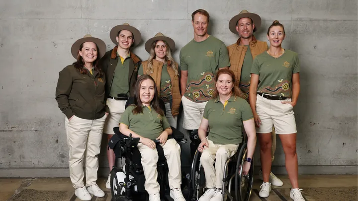 Australian Paralympic team members in uniform, two in wheelchairs, smiling indoors, concrete wall background.