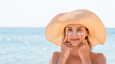 Woman in a sun hat applies sunscreen to her face at the beach.