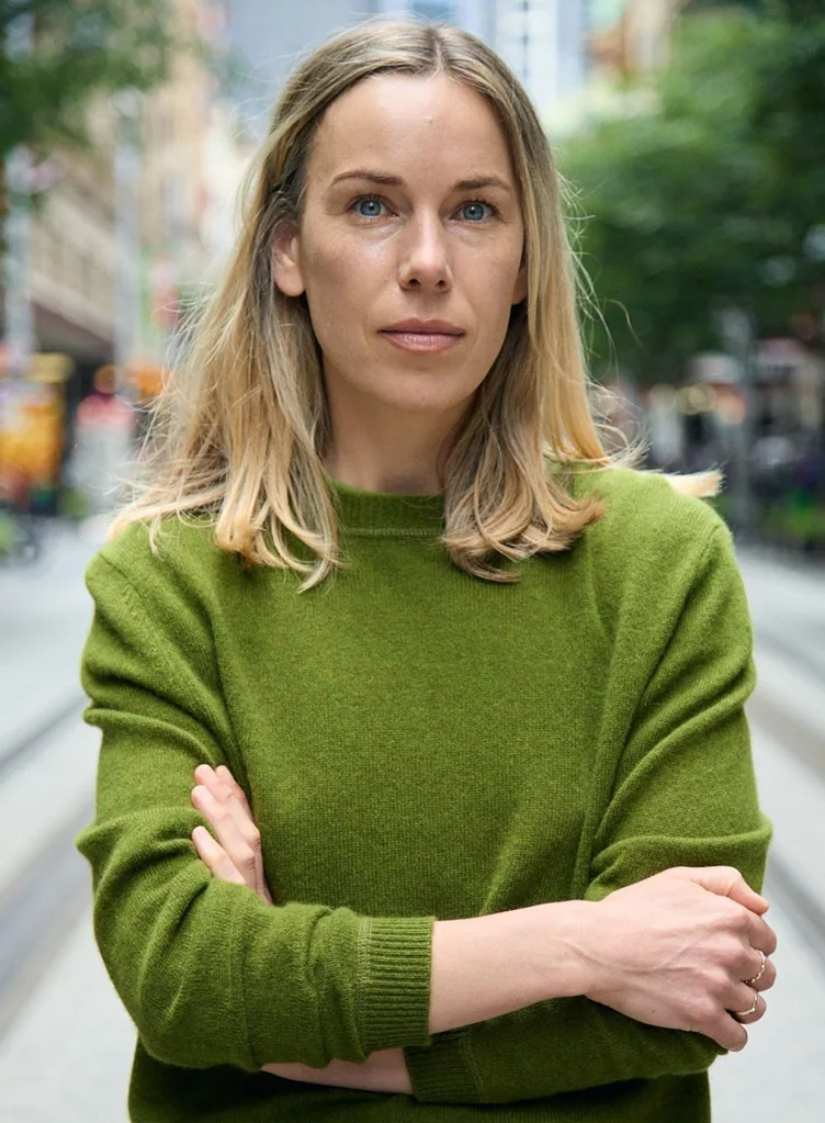 A portrait of Alexandra Gilbert, Head of Content at Prime Video, standing outdoors with her arms crossed. She has shoulder-length blonde hair and is wearing a moss-green crew neck cashmere jumper from the Jac + Jack AW26 collection. The city street background is softly blurred.