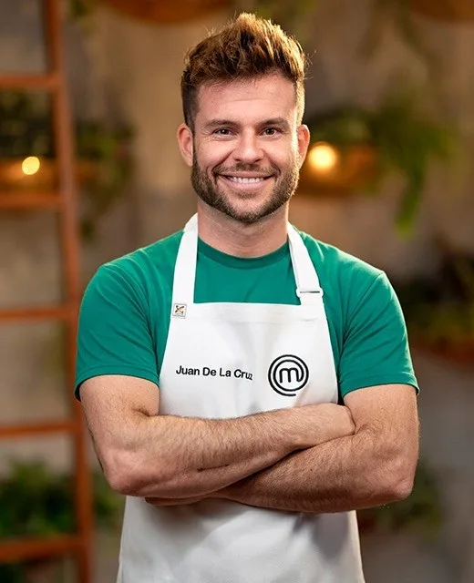 Contestant with brown hair wearing a "Juan De La Cruz" MasterChef apron, smiling with arms crossed in a kitchen setting.