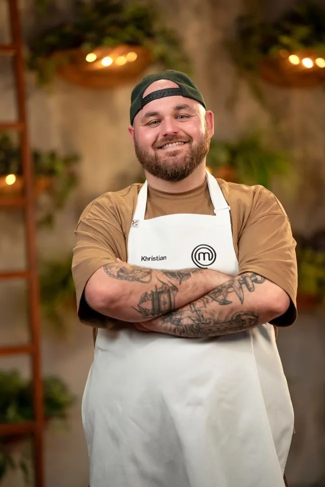 Man with a beard and tattoos in a MasterChef apron, smiling with arms crossed in a room with plants and warm lighting.