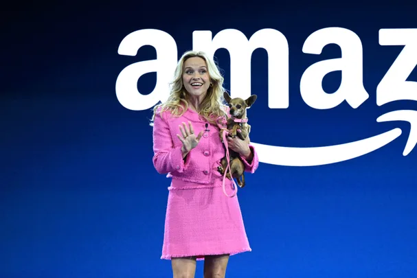 A woman in a pink outfit holds a small dog, standing in front of an Amazon logo on a blue background.