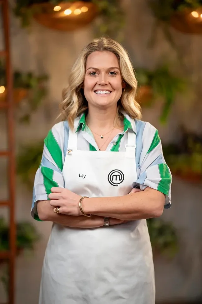 Female contestant named Lily wearing a "MasterChef" apron, smiling, arms crossed, in a bright kitchen setting.