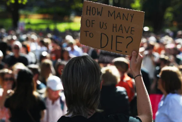 Person at protest holding a sign reading, "How many of us have to die?" in front of a large crowd.