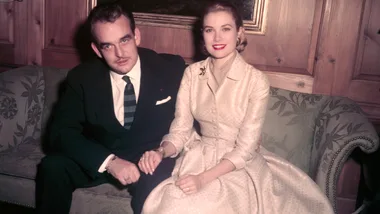 A man in a dark suit and a woman in a light dress sit on a floral sofa, holding hands and smiling at the camera.