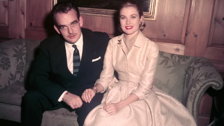A man in a dark suit and a woman in a light dress sit on a floral sofa, holding hands and smiling at the camera.