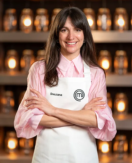 Woman with long hair and pink shirt wearing a "MasterChef" apron labeled "Snezana," standing with arms crossed, smiling.