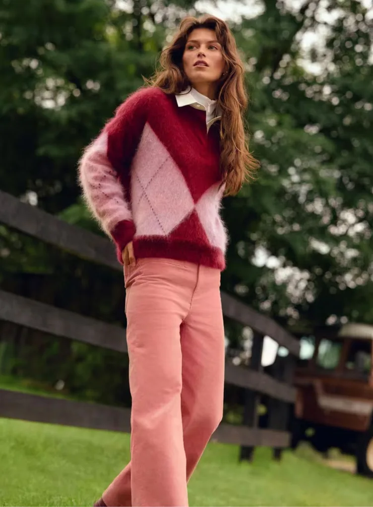 A model standing outdoors wearing the Sportscraft Tatiana Mohair Argyle Knit, a fuzzy, long-sleeved jumper featuring a bold pink and deep red diamond pattern. It is styled over a white collared shirt and paired with rose-pink wide-leg trousers. Best knitwear brands Australia article.