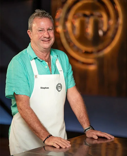 Man in a teal shirt and apron with "Stephen," standing in a kitchen studio with a MasterChef logo in the background.